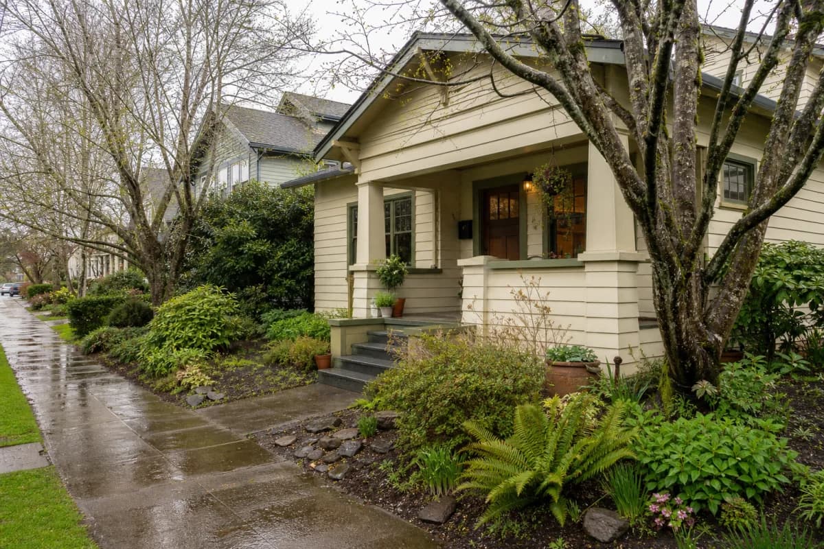 Portland Craftsman home with a covered porch, mature plants, and wet pavement